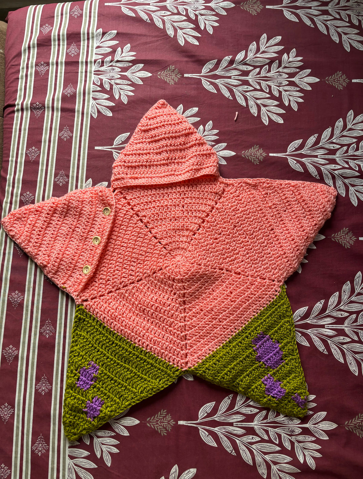A handmade crocheted hooded cape shaped like a star, featuring a coral pink top section with a hood and small buttons, and olive green lower sections