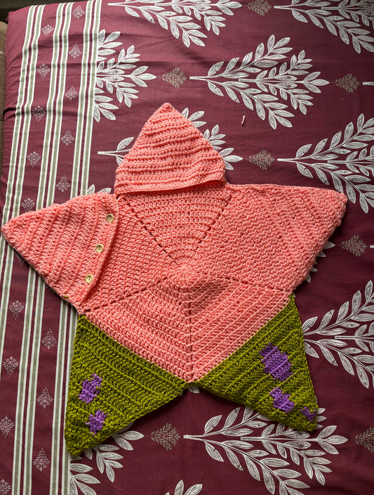 A handmade crocheted hooded cape shaped like a star, featuring a coral pink top section with a hood and small buttons, and olive green lower sections
